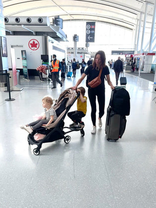  A mom with two young children at the airport. One child is riding on the stroller, while the other is walking beside her. The mom is carrying a small cross-body bag and pulling a suitcase